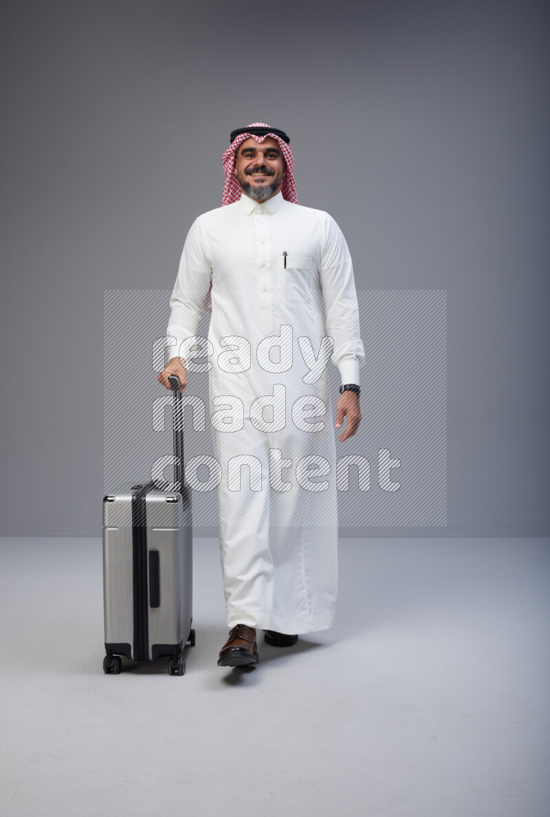 Saudi man wearing Thob and red Shomag standing holding Travel bag on Gray background