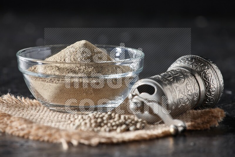 A glass bowl full of white pepper powder with white pepper beads on a burlap piece of fabric and a metal grinder on textured black flooring