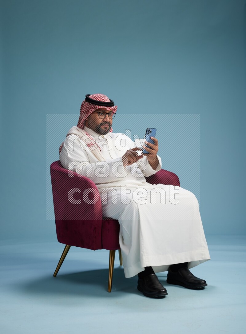 Saudi Man with shimag sitting on chair holding ATM card while talking on phone on blue background