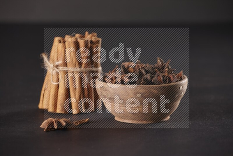 A stacked and bounded cinnamon sticks and a wooden bowl full of star anise on a black background