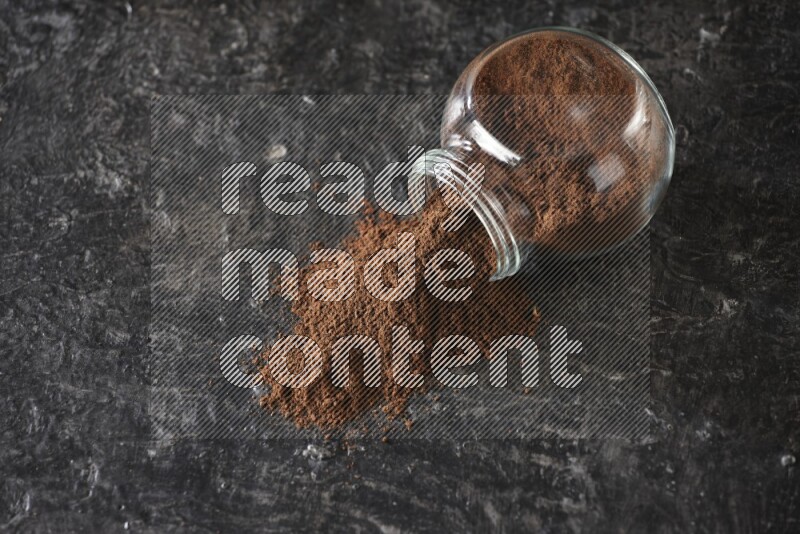 A flipped glass spice jar full of cloves powder and powder came out of it on textured black flooring