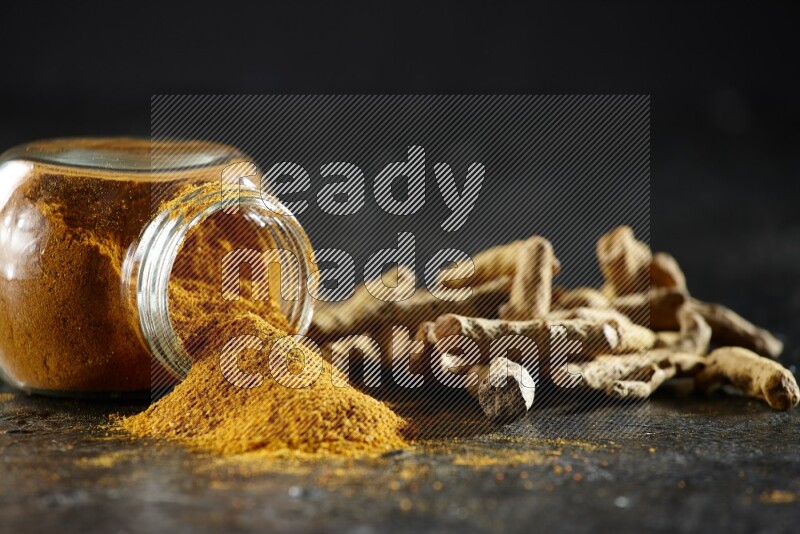 A flipped glass spice jar full of turmeric powder and powder spilled out of it with dried whole fingers on textured black flooring