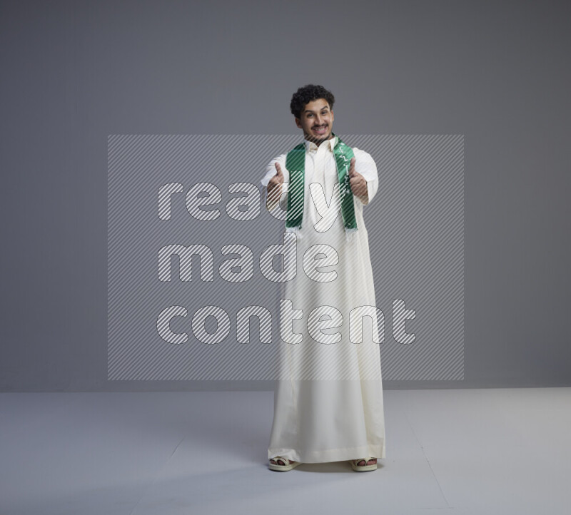 A Saudi man standing wearing thob and Saudi flag scarf on gray background