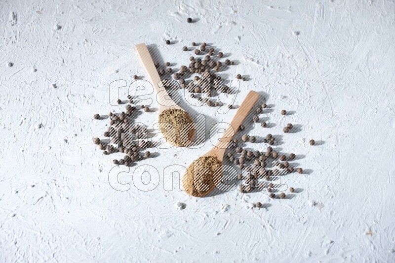 Wooden spoons full of allspice powder and allspice whole balls spreaded on a textured white flooring