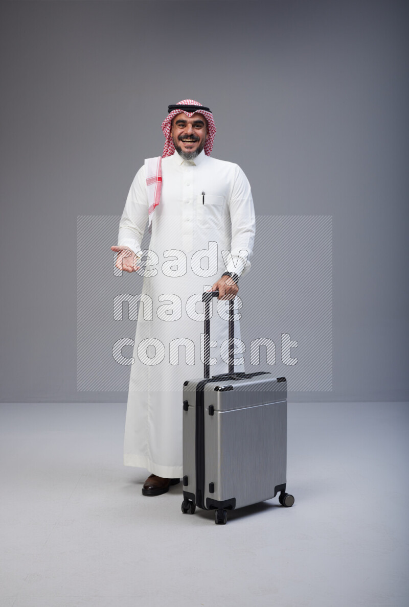 Saudi man wearing Thob and red Shomag standing holding Travel bag on Gray background