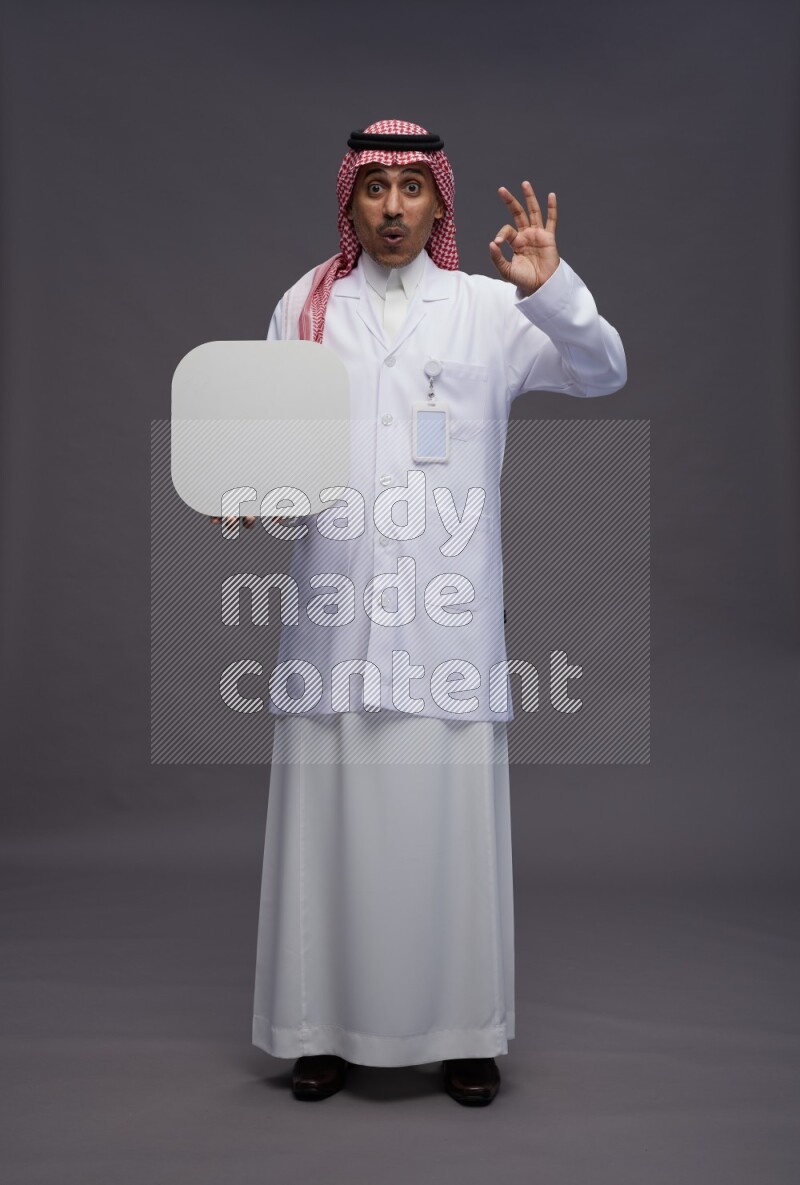 Saudi man wearing thob with lab coat and shomag with pocket employee badge standing holding social media sign on gray background