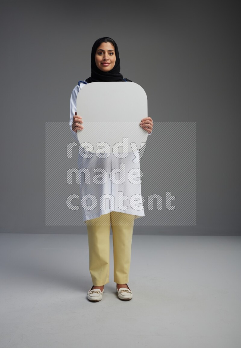 Saudi woman wearing lab coat with stethoscope standing holding social media sign on Gray background