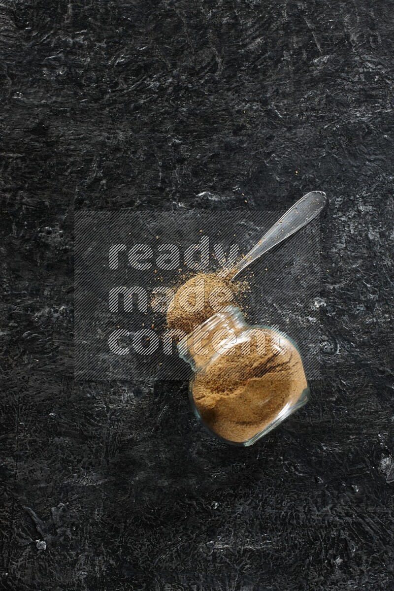 وعاء زجاجي للتوابل مقلوب وملعقة معدنية ممتلئة ببودرة الكمون والبودرة تناثرت منه على خلفية سوداء