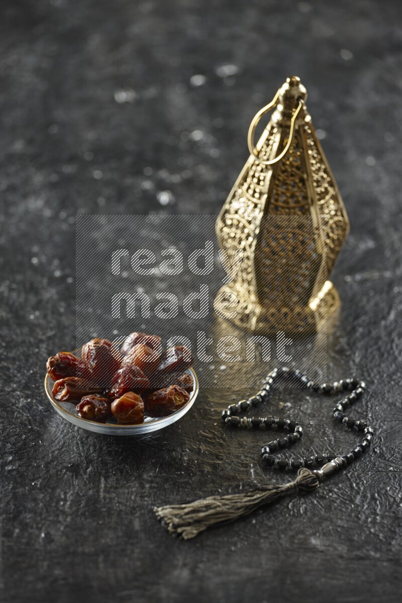 A golden lantern with different drinks, dates, nuts, prayer beads and quran on textured black background