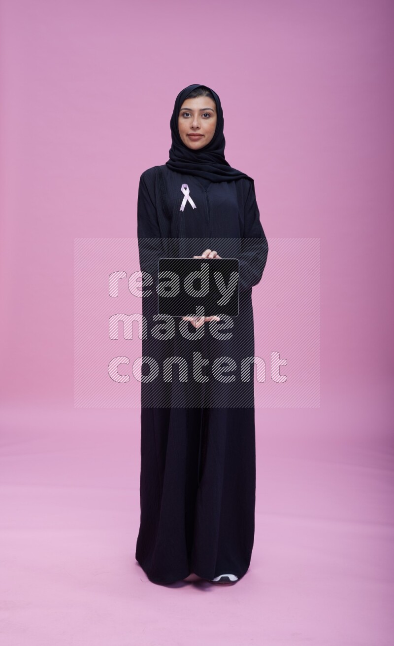 Saudi woman wearing pink ribbon on Abaya standing showing a tablet to camera on pink background