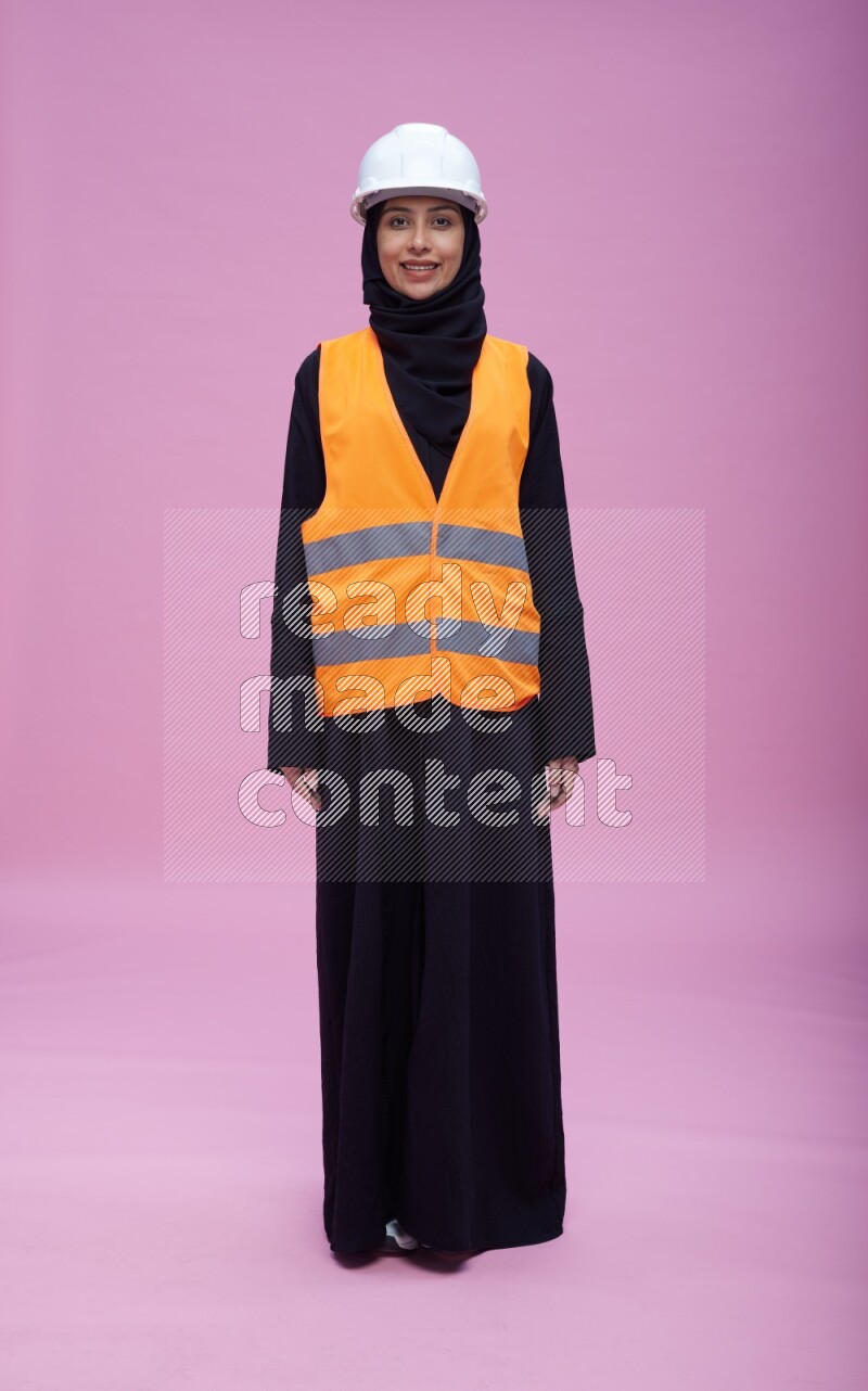 Saudi woman wearing Abaya with engineer vest and helmet standing interacting with the camera on pink background