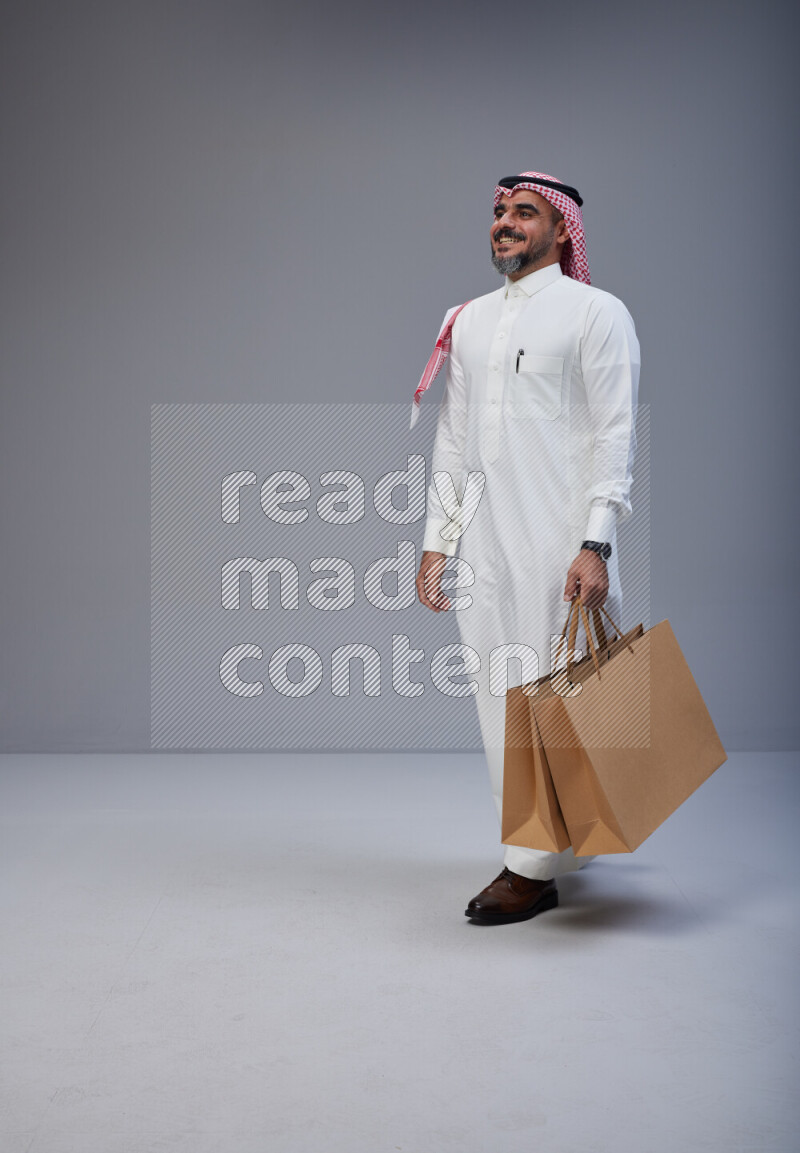Saudi man Wearing Thob and red Shomag standing holding shopping bag on Gray background