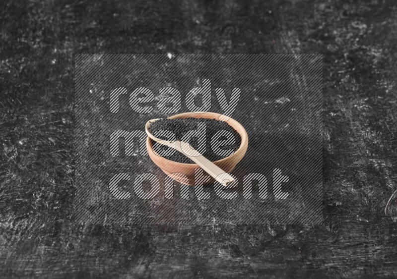 A wooden bowl full of black seeds with wooden spoon full of the seeds on it on a textured black flooring