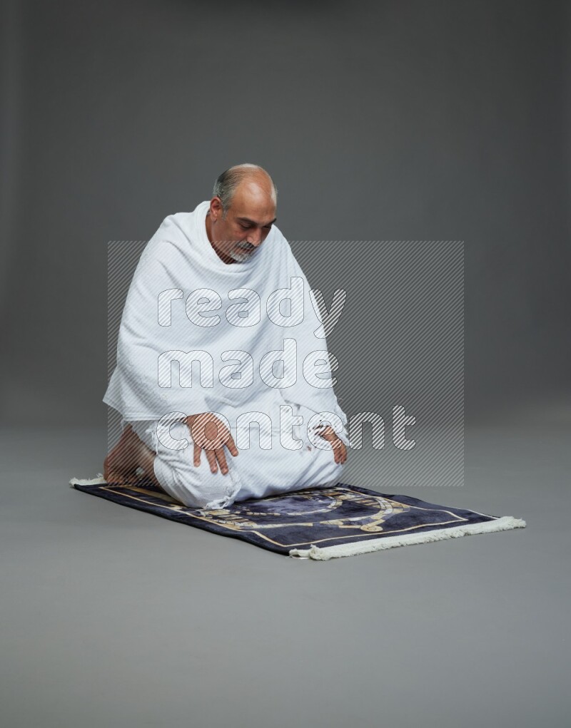 A man wearing Ehram sitting on prayer mat on gray background