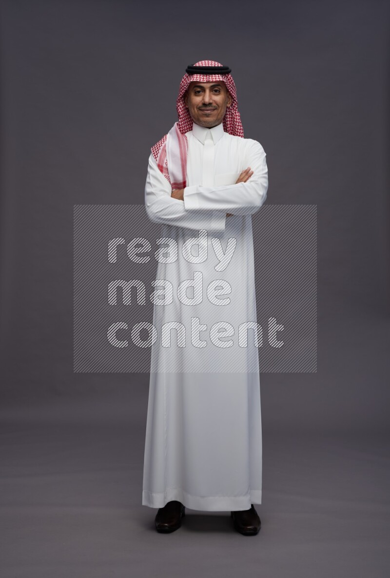 Saudi man wearing thob and shomag standing with crossed arms on gray background