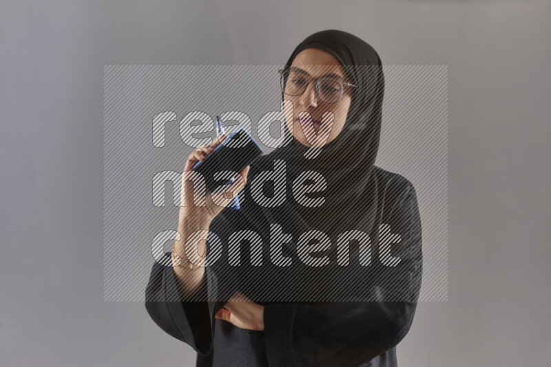 Woman in black abaya, hijab and glasses holding a marker pen to write on a transparent board with different reactions and expressions on grey background