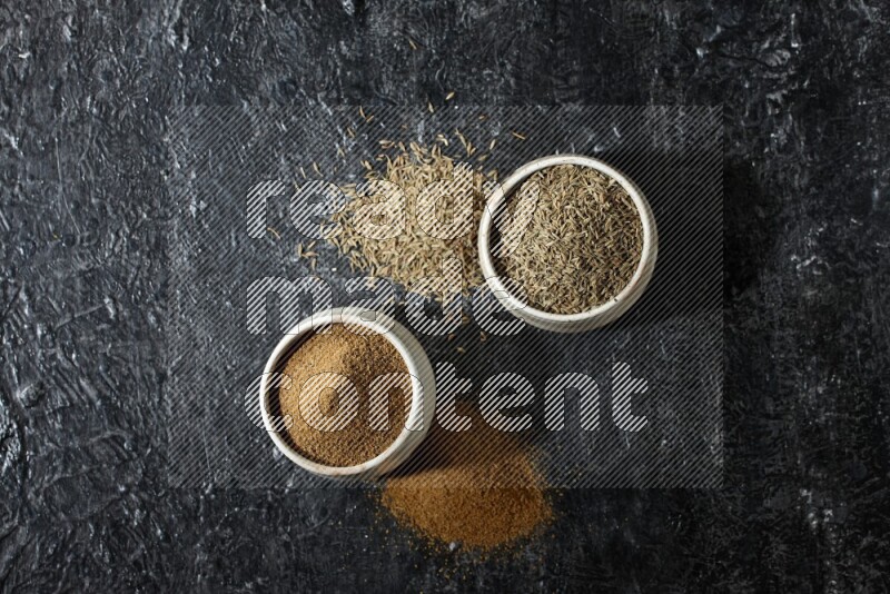 2 beige bowls, one full of cumin powder and the other full of seeds on a textured black flooring