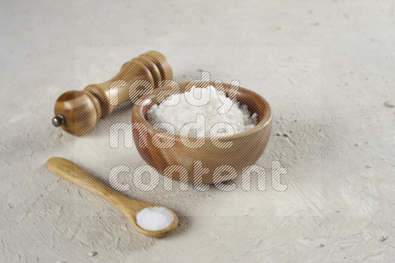 A wooden bowl and spoon filled with white sea salt and wooden grinder beside them on white background