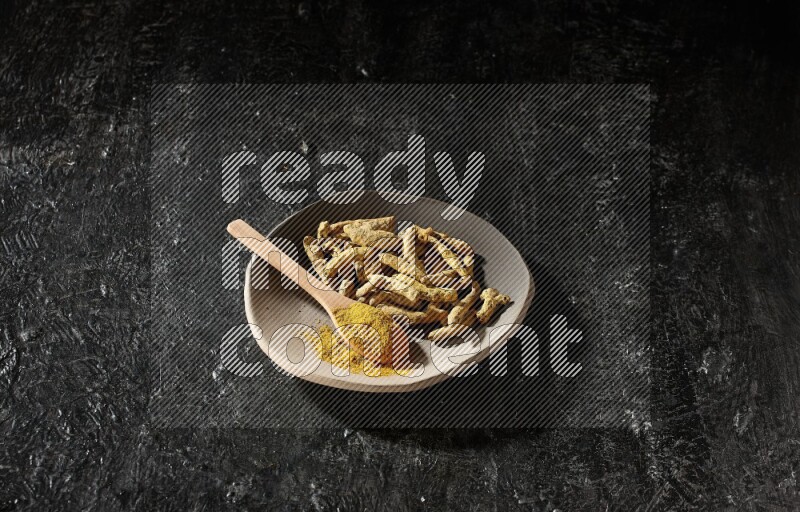 A plate filled with dried turmeric fingers and a wooden spoon full of turmeric powder on a textured black flooring