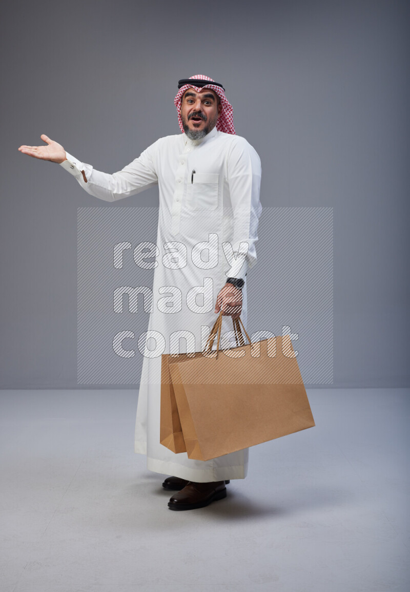Saudi man Wearing Thob and red Shomag standing holding shopping bag on Gray background