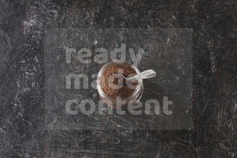 A glass jar full of cloves powder with a metal spoon on a textured black flooring