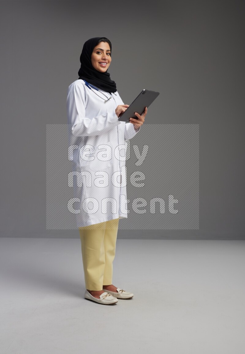 Saudi woman wearing lab coat with stethoscope standing working on tablet on Gray background