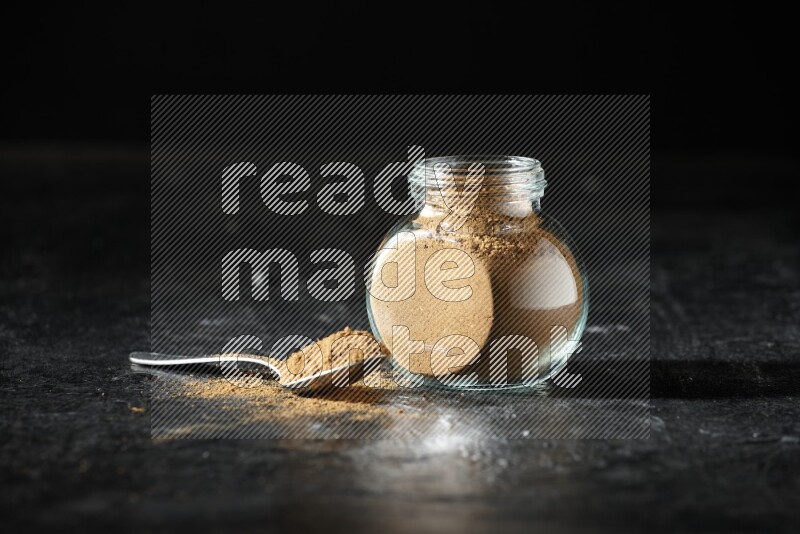 A glass spice jar and metal spoon full of allspice powder on a textured black flooring