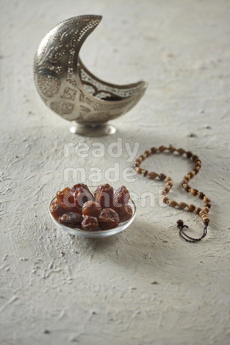 A silver lantern with different drinks, dates, nuts, prayer beads and quran on textured white background