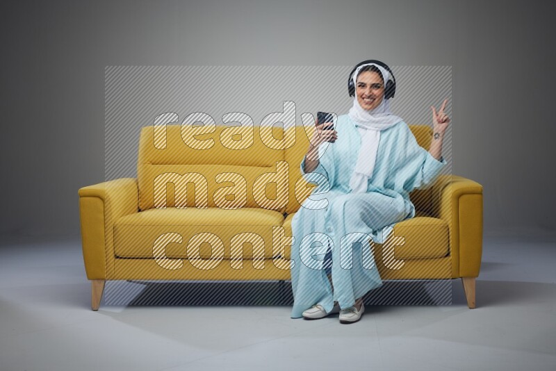 A Saudi woman wearing a light blue Abaya and a white head scarf sitting on a yellow sofa and holding an iPad while wearing headphone eye level on a grey background