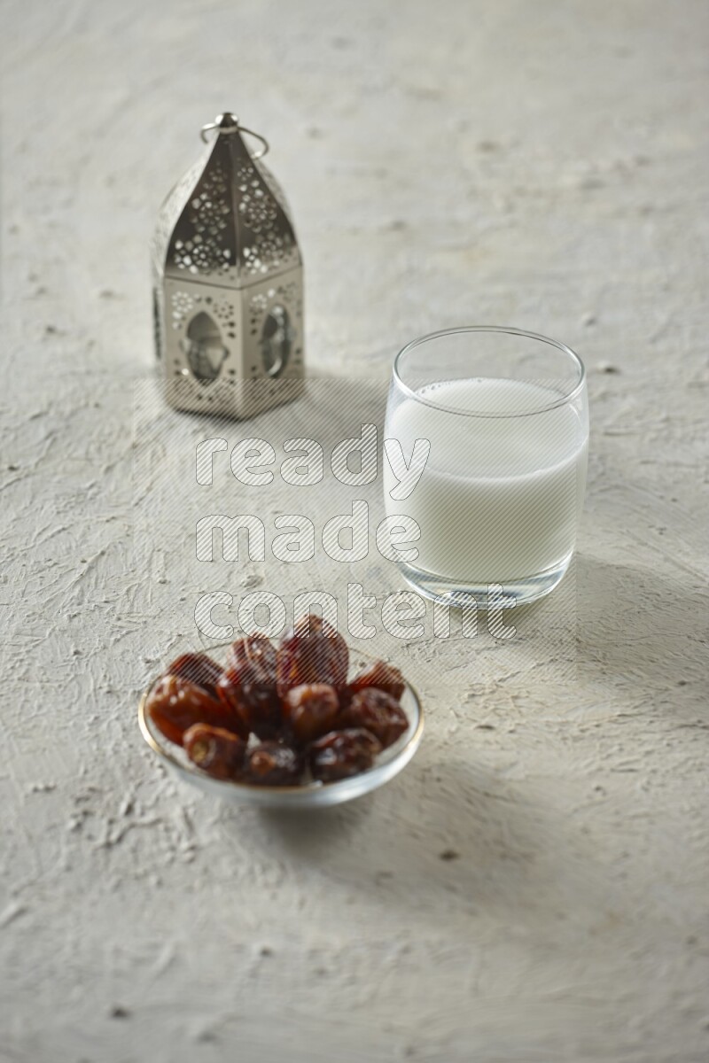 A silver lantern with different drinks, dates, nuts, prayer beads and quran on textured white background