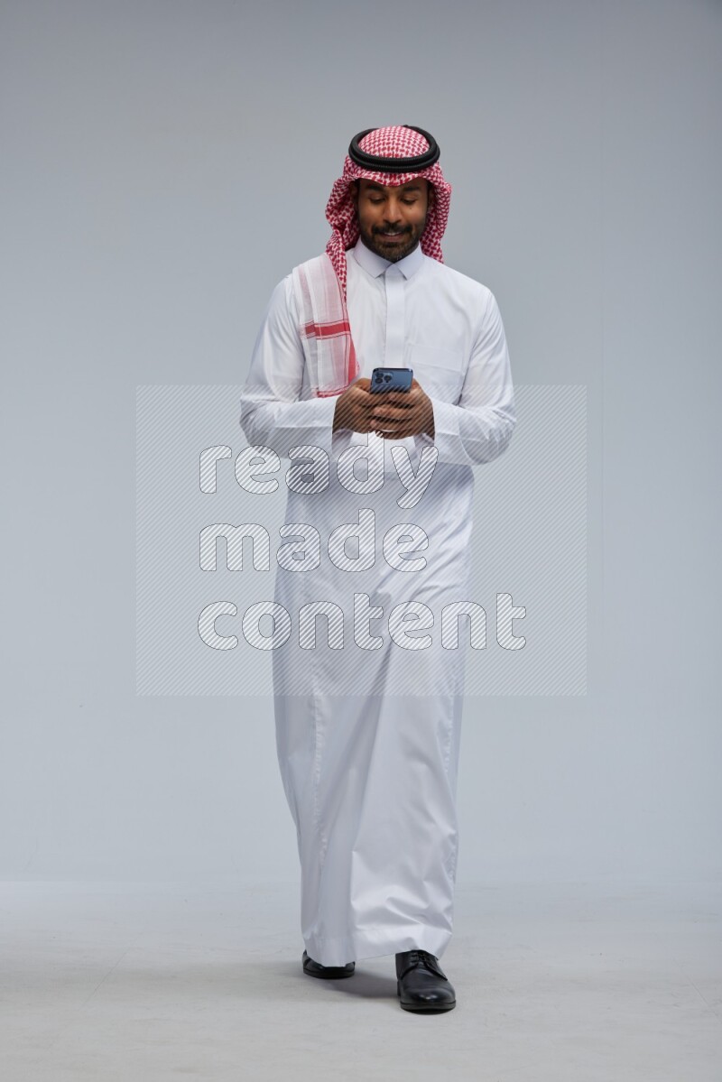 Saudi man Wearing Thob and shomag standing texting on phone on Gray background