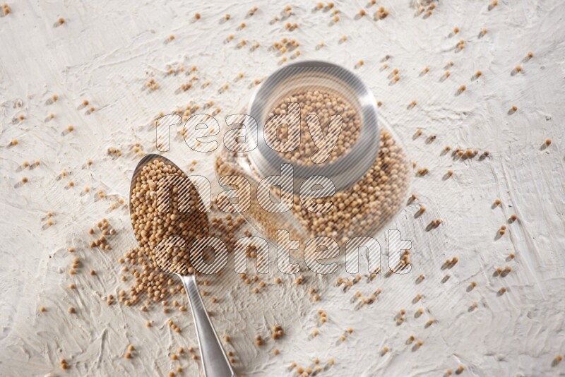 A glass spice jar and a metal spoon full of mustard seeds on a textured white flooring