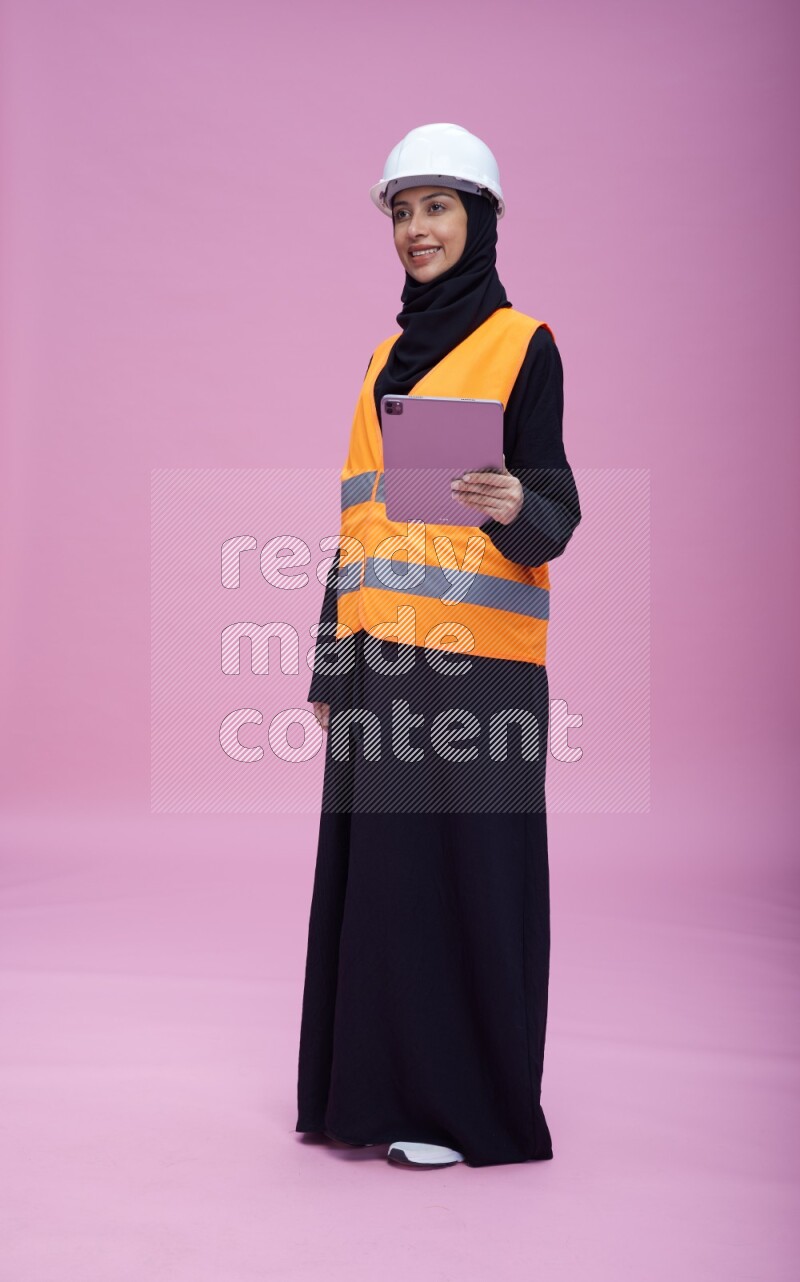Saudi woman wearing Abaya with engineer vest and helmet standing working on tablet on pink background