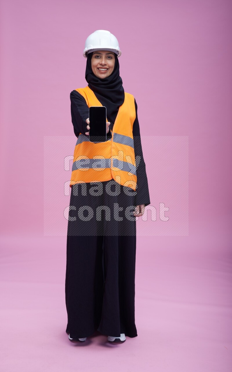 Saudi woman wearing Abaya with engineer vest and helmet standing showing phone to camera on pink background