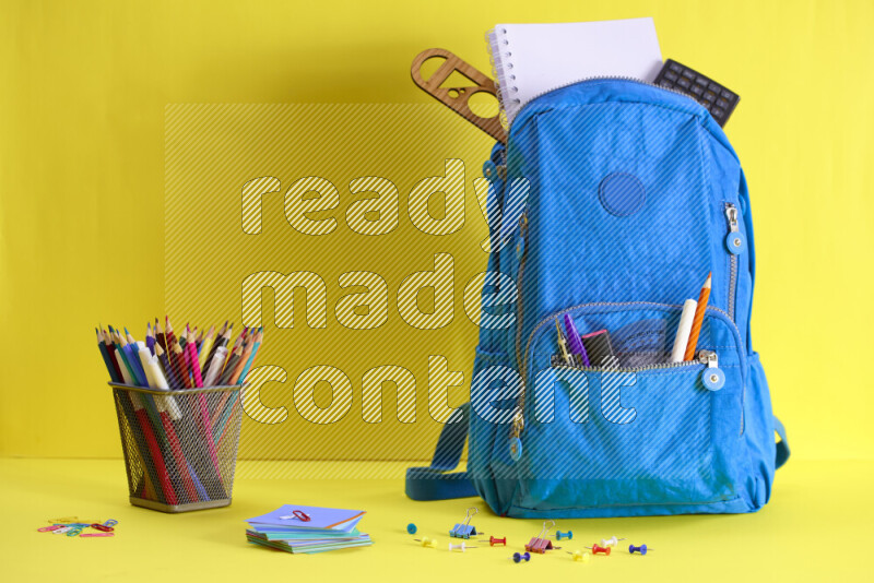 A school bag with assorted school supplies in and beside it on yellow background