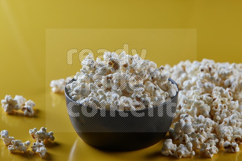 A black ceramic bowl full of popcorn with popcorn beside it on a yellow background in different angles