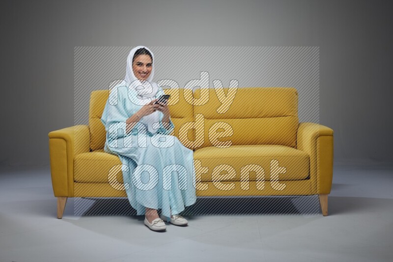A Saudi woman wearing a light blue Abaya and white head scarf sitting on a yellow sofa and using her phone eye level on a grey background