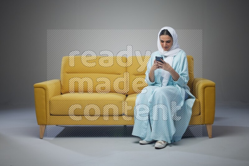 A Saudi woman wearing a light blue Abaya and white head scarf sitting on a yellow sofa and texting on her phone on a grey background