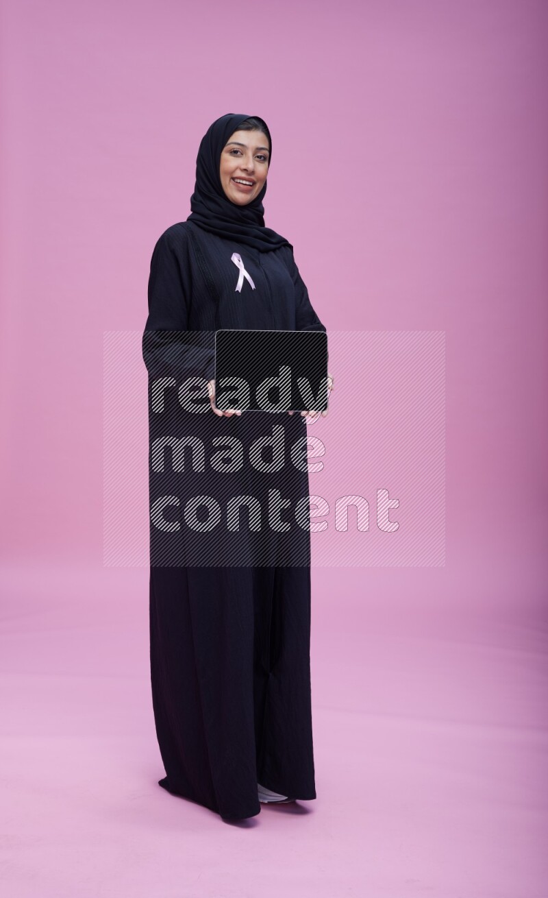Saudi woman wearing pink ribbon on Abaya standing showing a tablet to camera on pink background