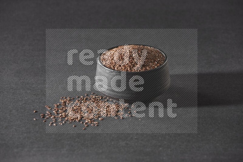 A black pottery bowl full of flaxseeds surrounded by the seeds on a black flooring