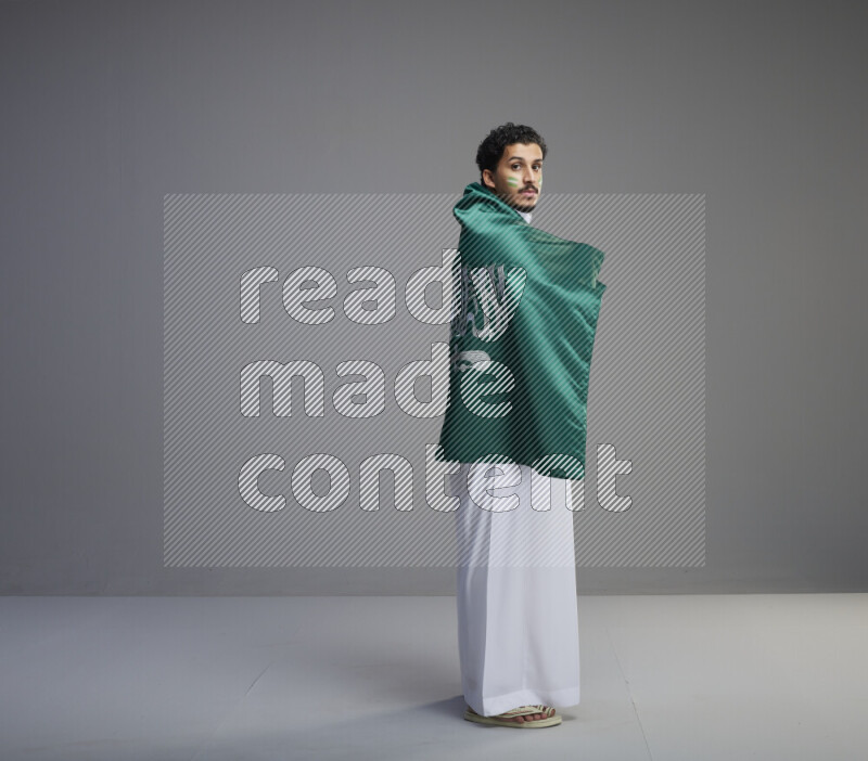 A Saudi man standing wearing thob with face painting wrapping big Saudi flag on gray background
