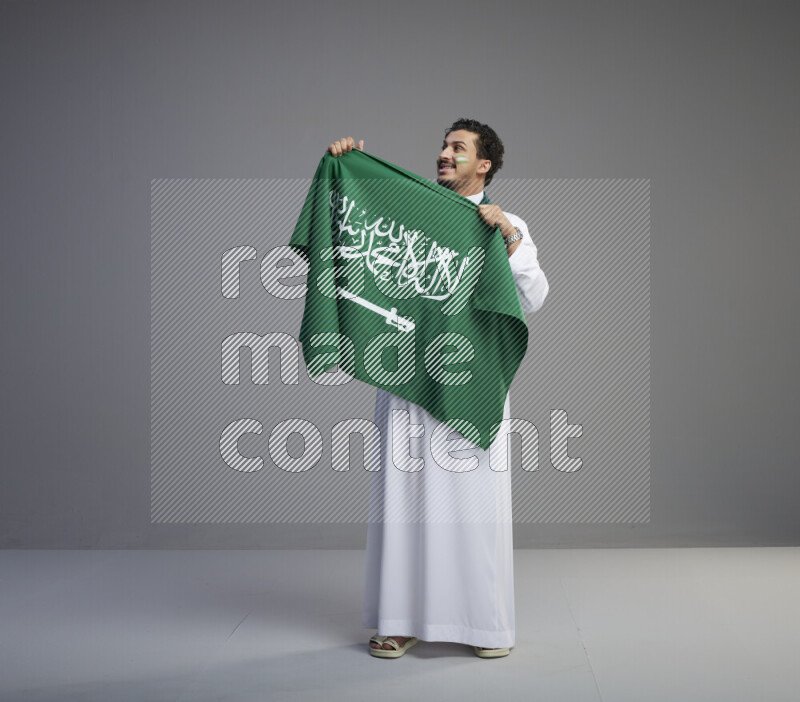 A Saudi man standing wearing thob with face painting wrapping Saudi flag scarf and holding big Saudi flag on gray background