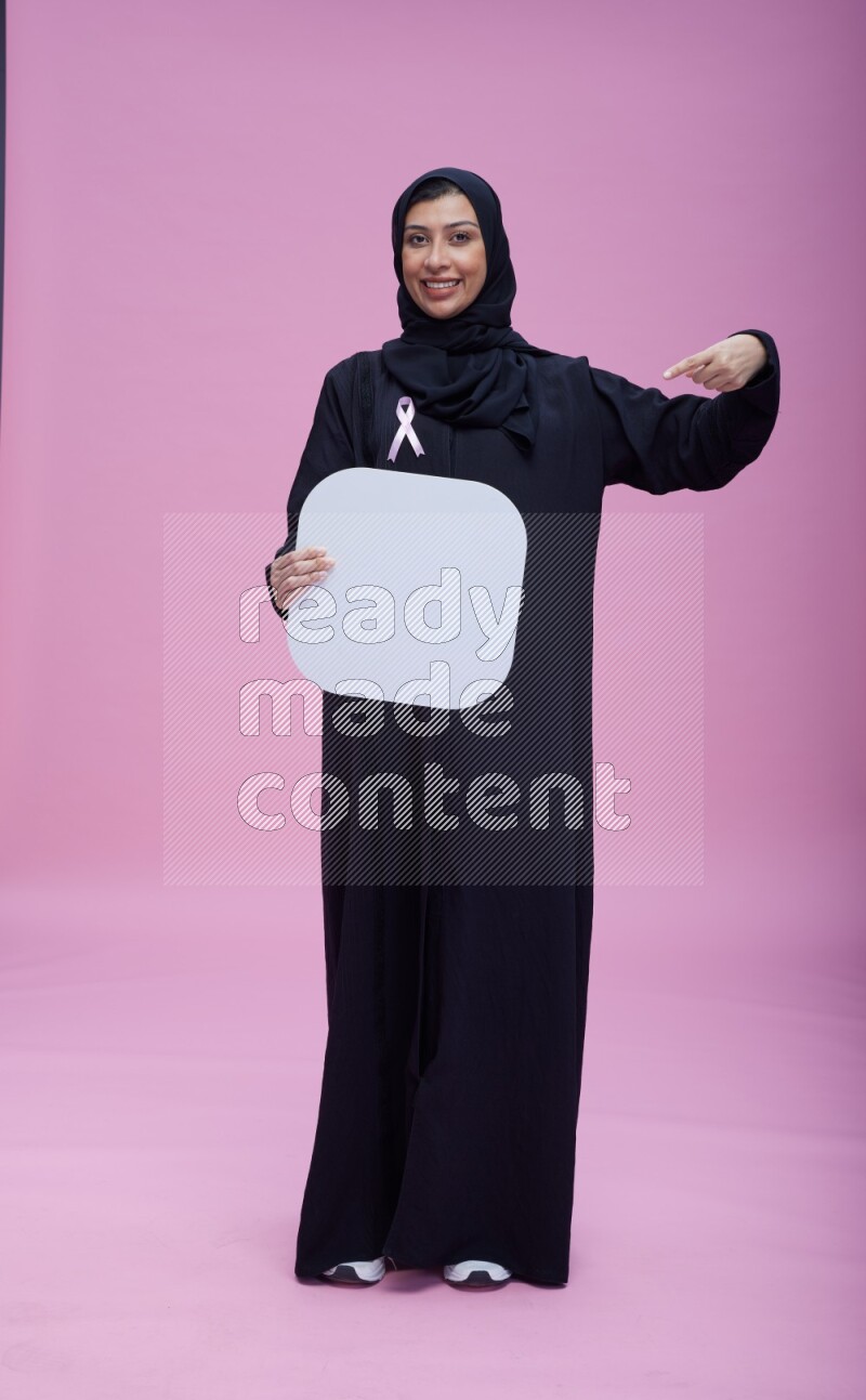 Saudi woman wearing pink ribbon on Abaya standing holding social media signs on pink background