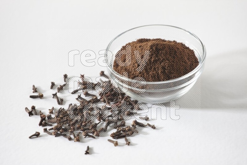 A glass bowl full of cloves powder and cloves grains spread on white flooring