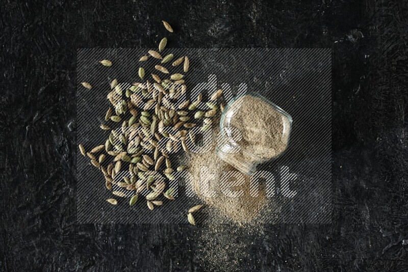 وعاء زجاجي للتوابل مقلوب وممتلئ ببودرة الهيل والبودرة تناثرت منه مع حبوب الهيل المتناثرة على خلفية سوداء