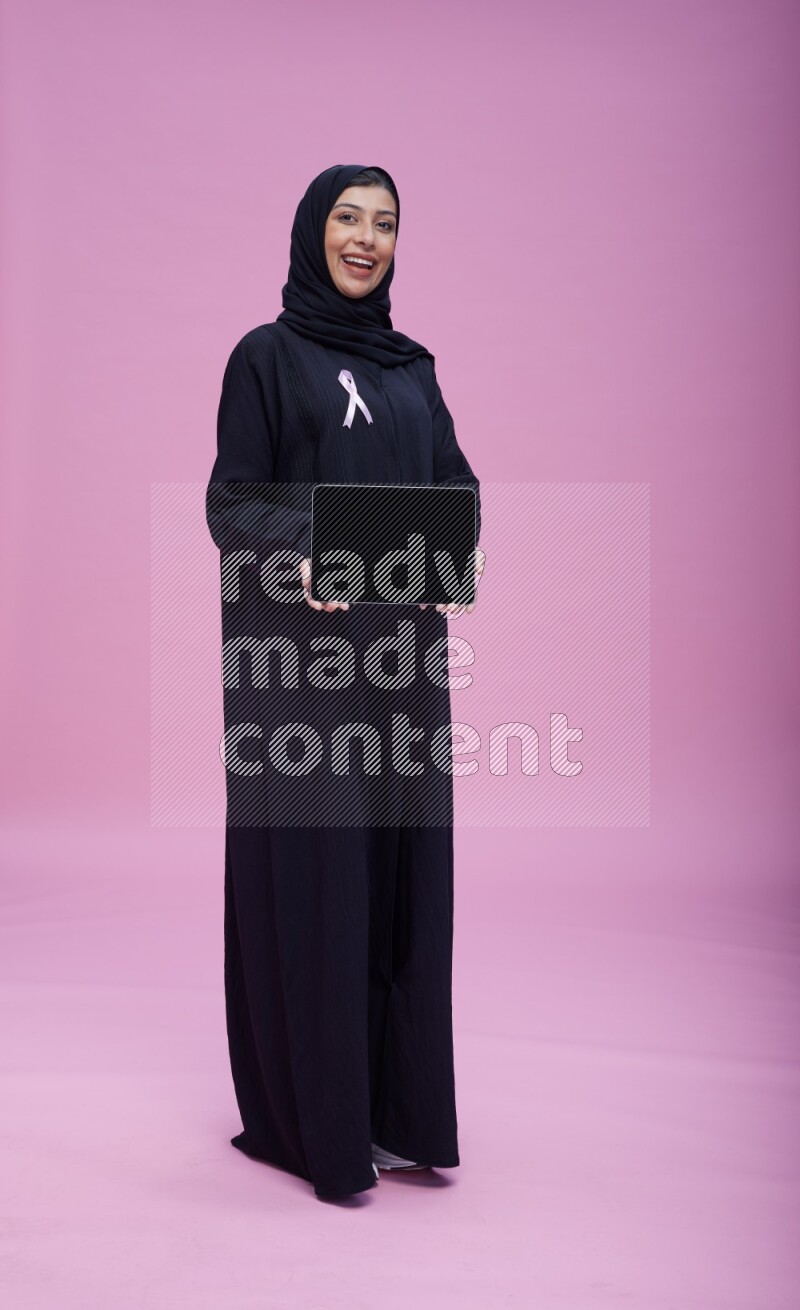 Saudi woman wearing pink ribbon on Abaya standing showing a tablet to camera on pink background