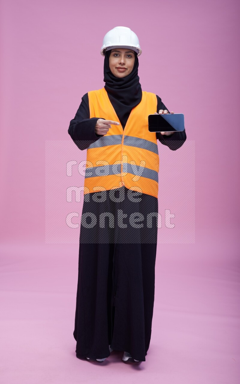 Saudi woman wearing Abaya with engineer vest and helmet standing showing phone to camera on pink background