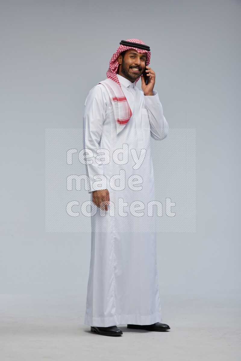 Saudi man Wearing Thob and shomag standing talking on phone on Gray background