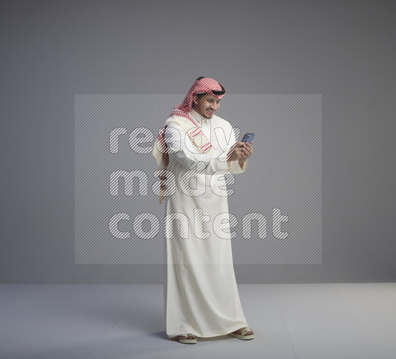 A  Saudi man standing wearing thob and red shomag texting on phone on gray background