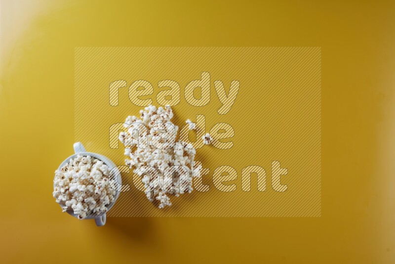 A white ceramic bowl full of popcorn with popcorn beside it on a yellow background in a top view shot